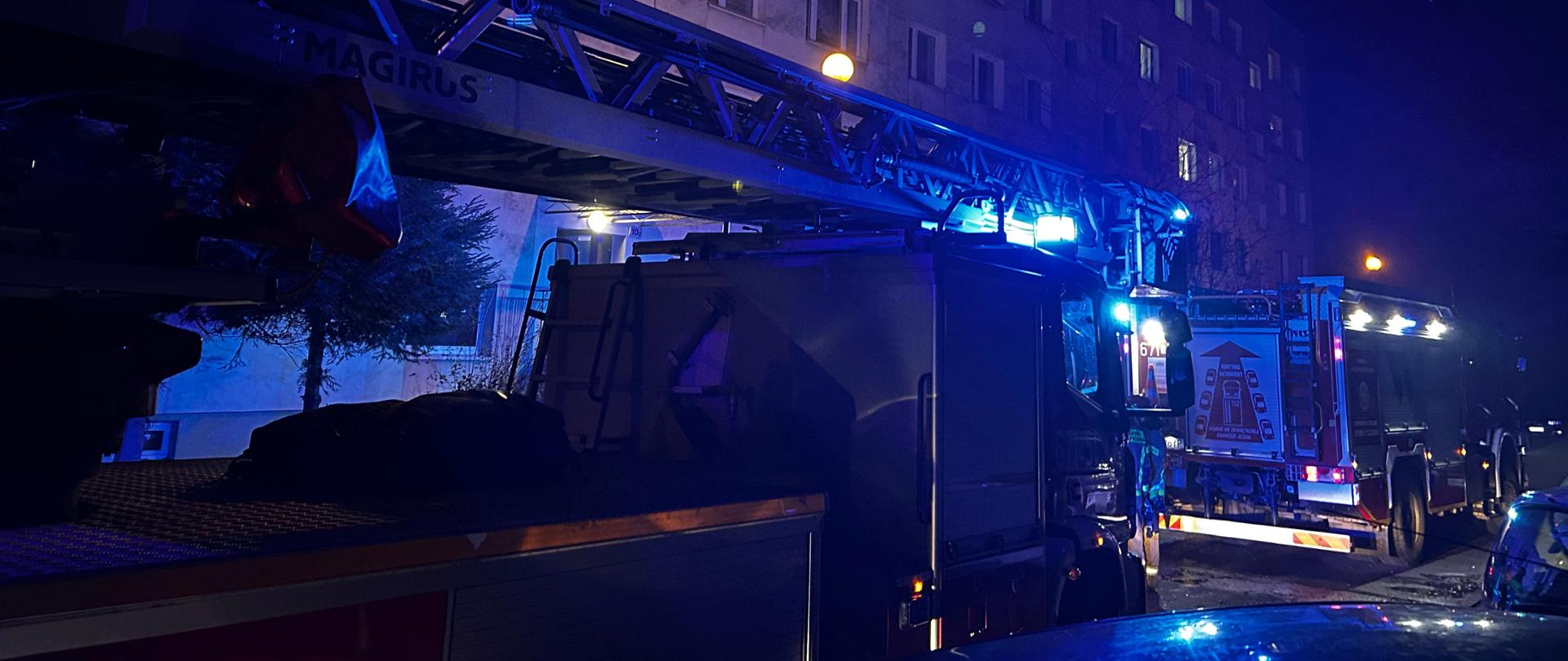 W nocy wozy strażackie z włączonymi niebieskimi światłami alarmowymi zaparkowane przed blokiem mieszkalnym podczas działań ratowniczych.