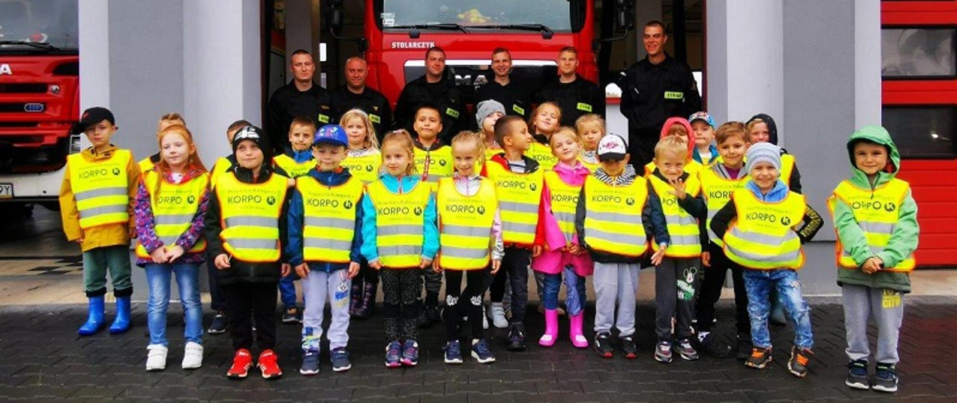 KP PSP w Białogardzie - wizyta dzieci z Przedszkola Miejskiego nr 2 w Białogardzie. Prewencja społeczna - akcja ”Kręci mnie bezpieczeństwo”. Zdjęcie grupowe dzieci i strażaków przed garażami samochodów strażackich