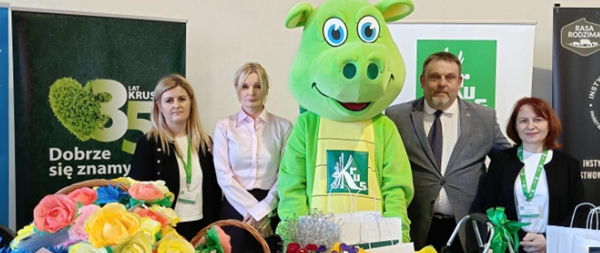 Przy stoisku informacyjno-prewencyjnym KRUS, w centrum stoi maskotka KRUS – osoba w zielonym pluszowym stroju smoka z logo KRUS. Obok niej stoją cztery osoby – trzy kobiety i jeden mężczyzna. Przed nimi widoczna trybunka reklamowa z logo KRUS, materiały prewencyjne, baner jubileuszowy „35 lat KRUS” oraz dekoracje kwiatowe.