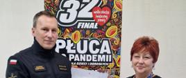 Strażak przekazujący płytkę z wizerunkiem św. Floriana wolontariuszowi WOŚP, w tle plakat 32 finału WOŚP