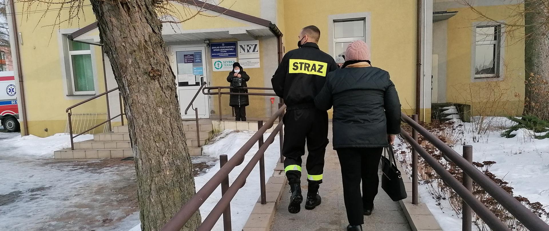 Strażak prowadzi pod rękę osobę starszą do punktu szczepień idąc podjazdem dla osób niepełnosprawnych. W tle widoczny jest punkt szczepień. 