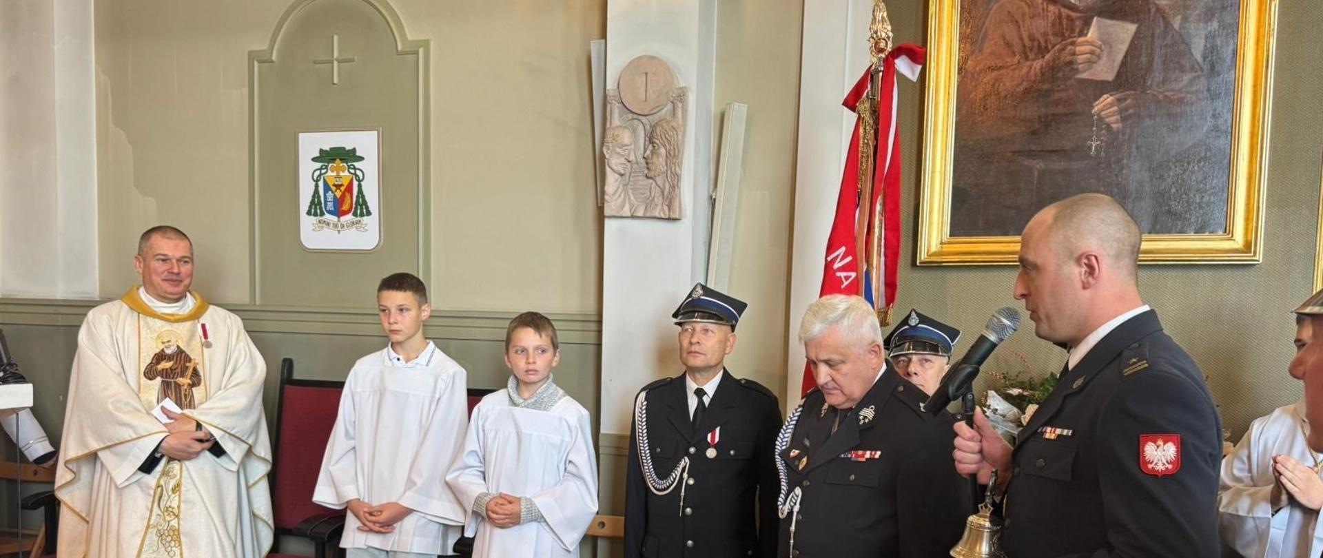 Kapelan strażaków, ministranci strażacy w tym Komendant Powiatowy trzymający prezent na pożegnanie kapelana