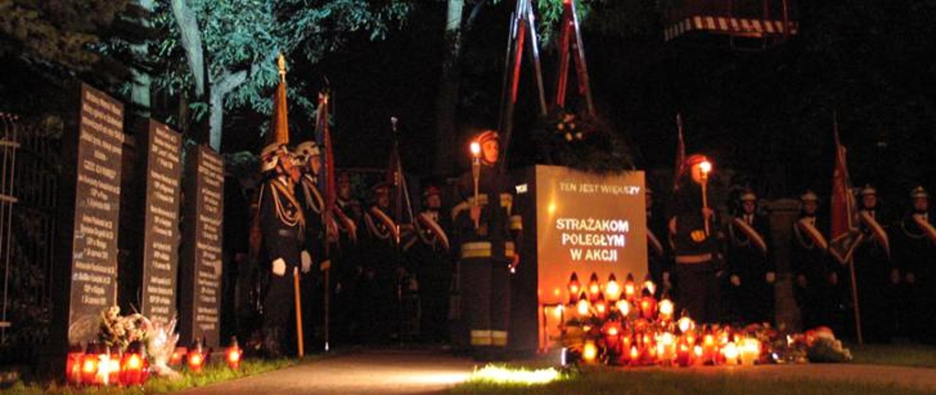 Pomnik z napisem "Strażakom poległym w akcji", dookoła zgromadzeni strażacy. Przed pomnikiem stoją zapalone znicze.