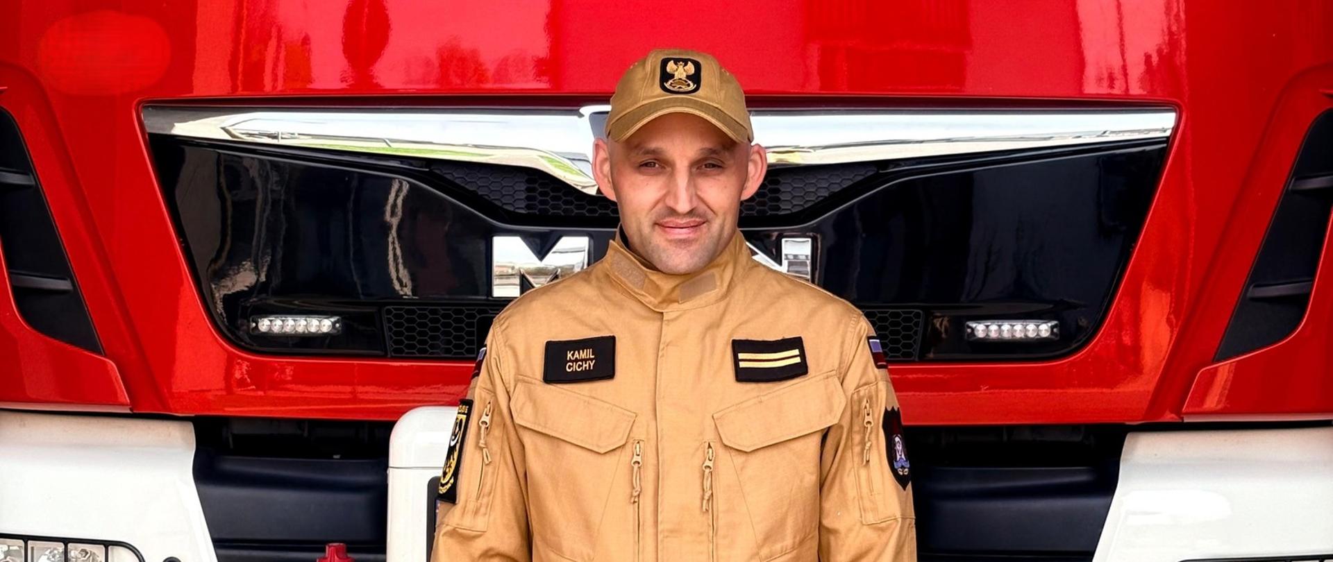 Portret strażaka w piaskowym mundurze służbowym i czapce z daszkiem z godłem, stojącego na tle przodu czerwonego samochodu ratowniczo-gaśniczego marki MAN. Mężczyzna patrzy w stronę obiektywu i lekko się uśmiecha. Na jego mundurze widoczna jest naszywka z imieniem i nazwiskiem „KAMIL CICHY” oraz patka ze stopniem służbowym (dwie poziome belki).