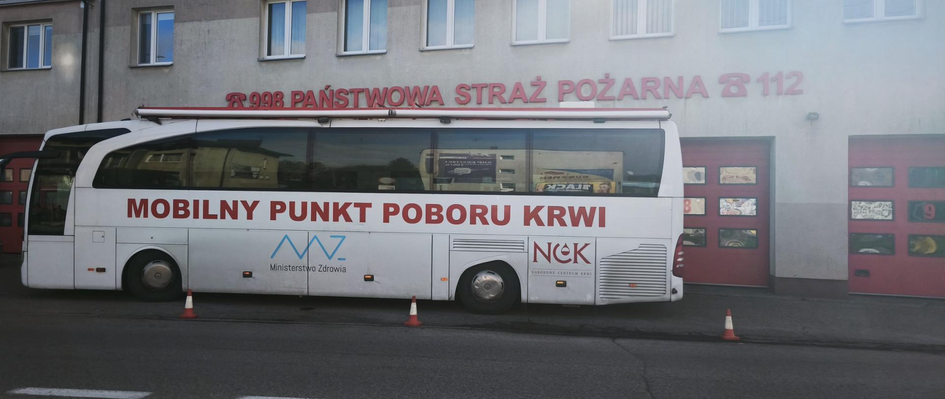 Na zdjęciu widać duży biały autobus zaparkowany przed budynkiem straży pożarnej. Na autobusie widnieje napis "MOBILNY PUNKT POBORU KRWI" oraz logo Ministerstwa Zdrowia (MZ) i Narodowego Centrum Krwi (NCK).
