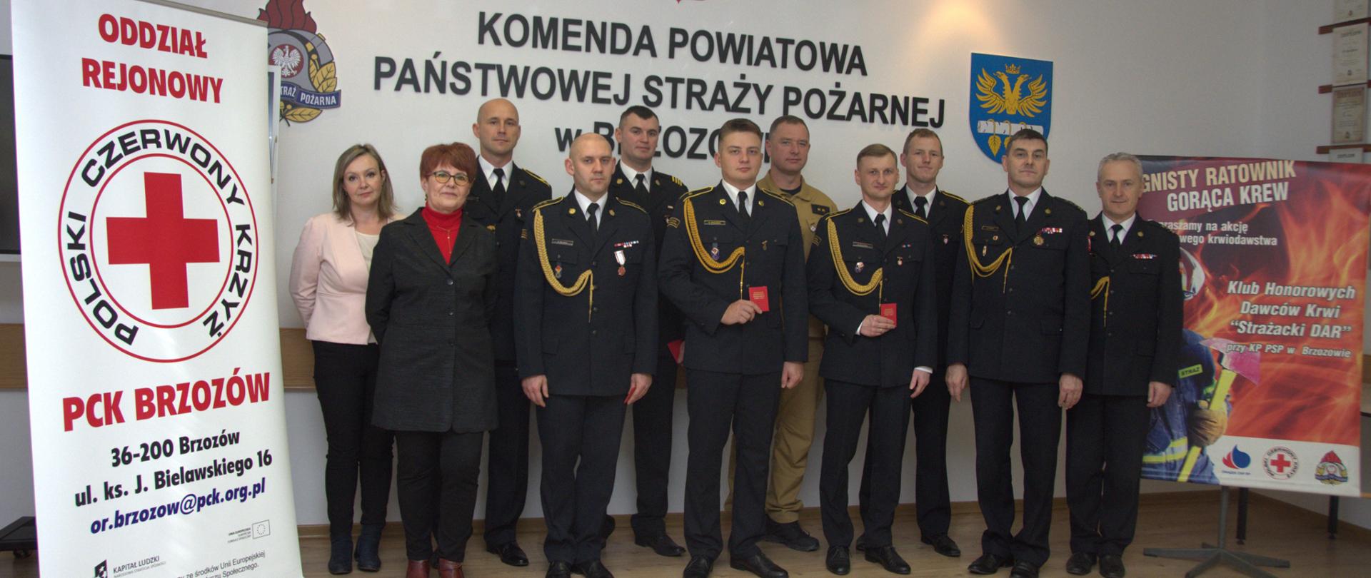 Kolorowa fotografia wykonana w świetlicy Komendy Powiatowej PSP w Brzozowie. Przedstawia grupę strażaków w mundurach wyjściowych i osoby cywilne po wręczeniu odznaczeń Honorowych Dawców Krwi.