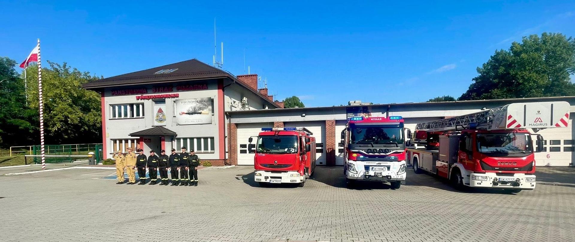 Przed budynkiem Komendy Miejskiej PSP w Świętochłowicach stoi w postawie zasadniczej ośmiu strażaków. Jeden z nich oddaje honor. Po prawej stronie widać zaparkowane trzy samochody pożarnicze. 