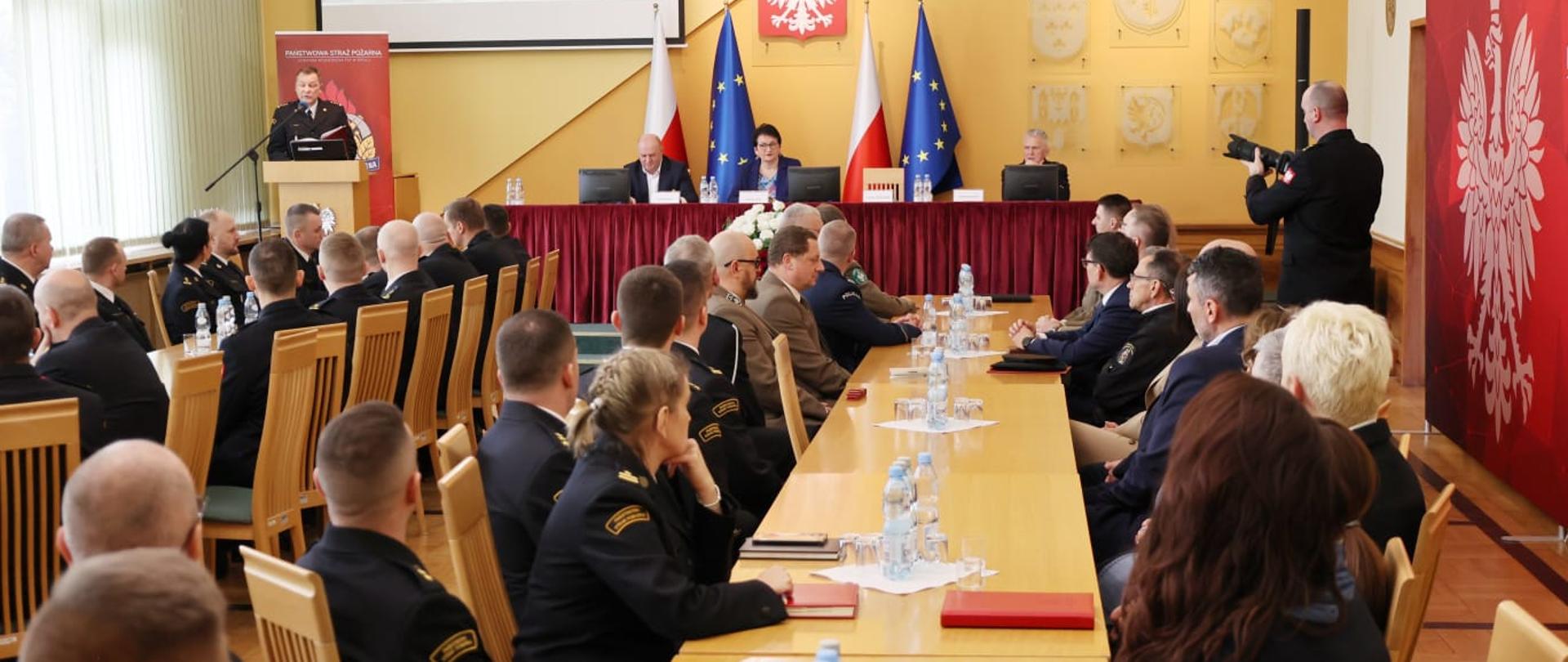 Narada roczna Kadry Kierowniczej Państwowej Straży Pożarnej województwa opolskiego. Na zdjęciu widać siedzących przy stołach uczestników. W tle widać Wojewodę Opolskiego oraz Marszałka Województwa Opolskiego oraz przemawiającego w Opolskiego Komendanta Wojewódzkiego Państwowej Straży Pożarnej, Pana Pawła Kielara. 