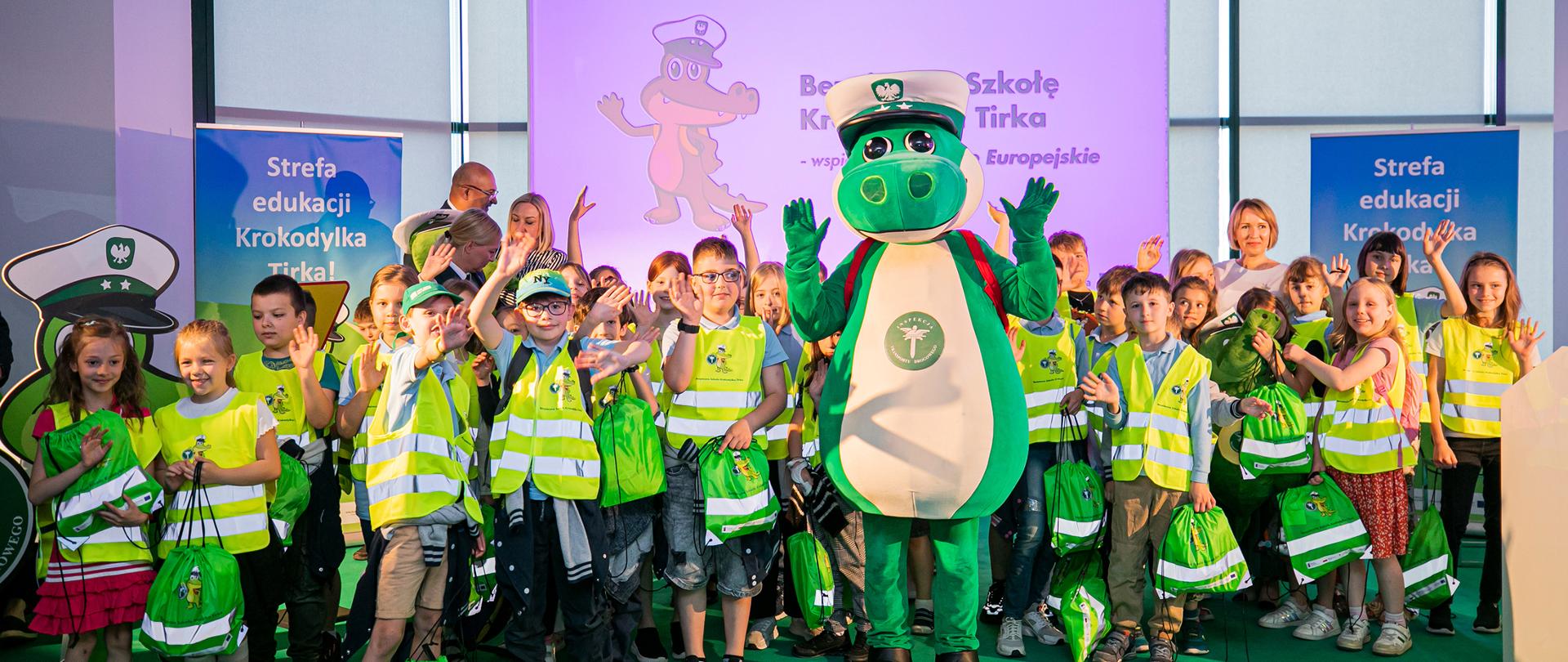 Dzieci stoją na scenie - na zielonej wykładzinie. W środku między nimi stoi maskotka Krokodylek Tirek. Dzieci machają, mają odblaskowe kamizelki i plecaki z wizerunkiem Krokodylka Tirka. Z tyły ścianki z napisem Strefa edukacji Krokodylka Tirka!