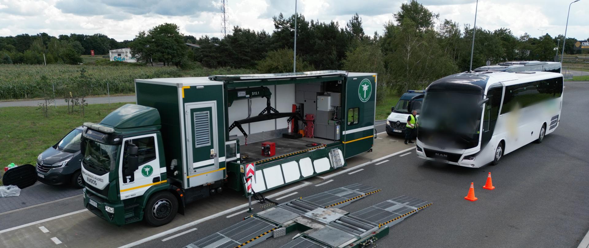Zdjęcie z szerokiej perspektywy przedstawia zieloną ciężarówkę z rozłożonym sprzętem kontrolnym. Obok niej stoi biały autokar. Na ziemi widać rampy do pomiarów, a w pobliżu stoją pomarańczowe pachołki. W tle widać inne pojazdy i teren zielony.