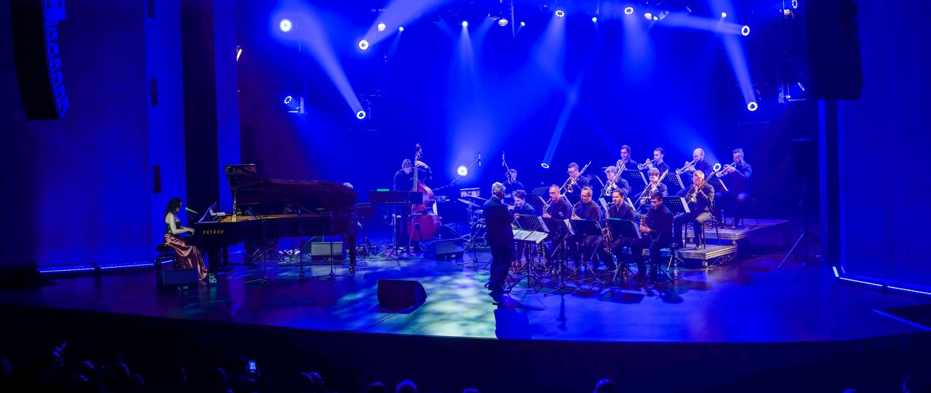 Zdjęcie Big bandu z Iloną Damięcką i Krzysztofem Pydyńskim. Zespół siedzi na scenie DK Oskard z instrumentami.