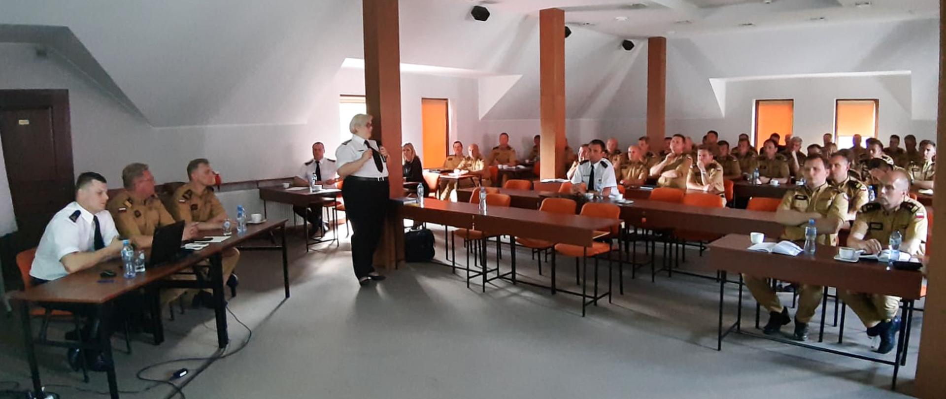 sala konferencyjna na poddaszu, w sali przy birkach siedzą strażacy czworo w białych koszulach pozostali w piaskowym umundurowaniu