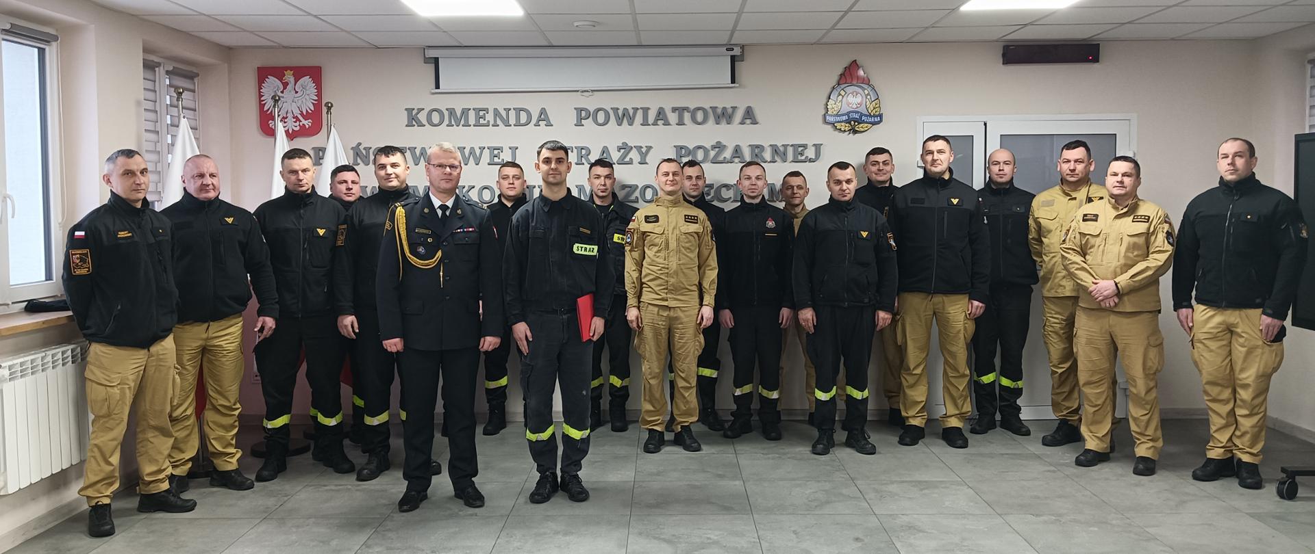 Wspólne zdjęcie funkcjonariuszy z Zastępcą Komendanta Powiatowego PSP w Makowie Mazowieckim, z tle jest ściana z napisem "Komenda Powiatowa Państwowej Straży Pożarnej w Makowie Mazowieckim".
