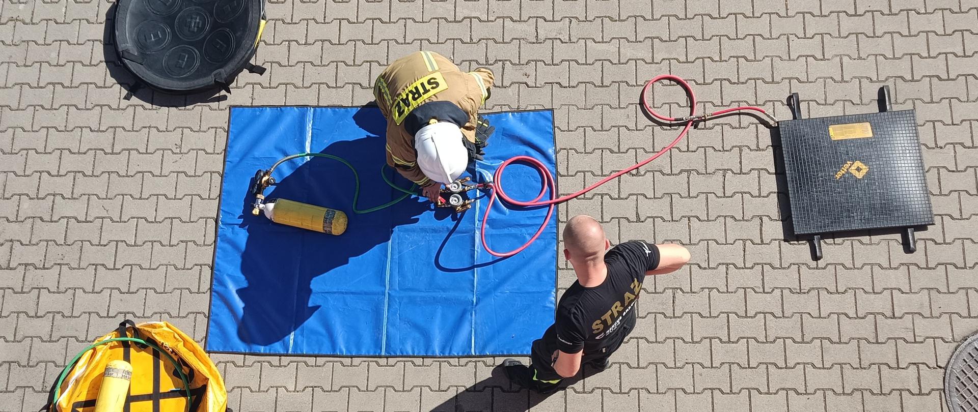 Na zdjęciu uczestnik szkolenia kierowców-konserwatorów sprzętu ratowniczego OSP, który odbył się w Komendzie Powiatowej PSP w Pabianicach. Na zdjęciu jeden z kursantów sprawia sprzęt pneumatyczny podczas egzaminu praktycznego. Obok niebieskiej maty stoi egzaminator (strażak KP PSP w Pabianicach). Dookoła rozłożony sprzęt strażacki.