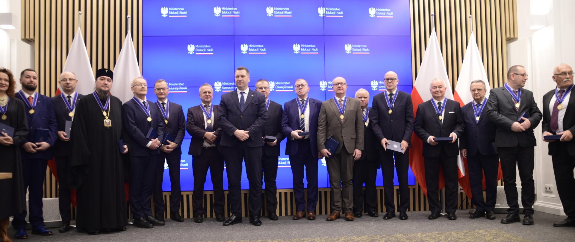 Na zdjęciu minister Czarnek z kilkunastoma osobami (w tym osoba duchowna) ubranymi w uroczyste stroje, na szyjach zebranych wiszą medale. W tle ekran z grafiką: na niebieskim tle widnieją logotypy MEiN, po bokach polskie flagi.