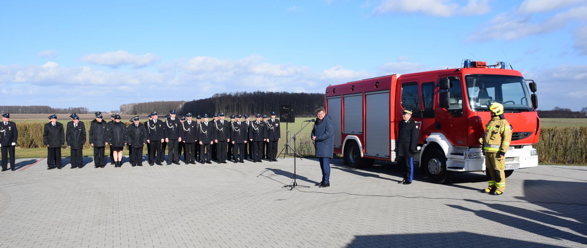 Na zdjęciu po prawej stronie samochód pożarniczy koloru czerwonego. W tle pododdział strażaków w mundurach wyjściowych koloru granatowego. 