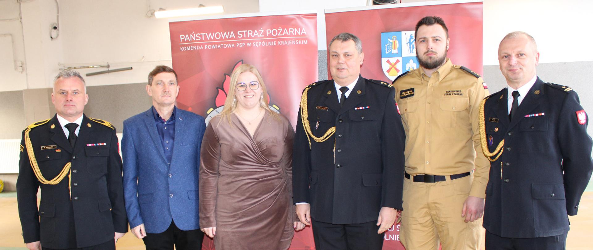 Komendanci powiatowi w mundurach wyjściowych przewodniczący związków zawodowych w mundurach wyjściowym i służbowym oraz pracownicy cywilni w ubraniach okazjonalnych na tle baneru w pomieszczeniach garażowych komendy powiatowej w sępólnie krajeńskim pozują do zdjęcia