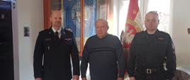 Komendant powiatowy oraz zastępca komendanta stoją po obu stronach druha OSP, który w lewej dłoni trzyma decyzję o przyznaniu świadczenia ratowniczego. W tle sztandar KP PSP w Żyrardowie.