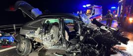 Rozbity samochód osobowy na pasie drogi krajowej nr 7. Na drugim palnie karetka pogotowia oraz pojazdy pożarnicze z włączoną sygnalizacją świetlną. 