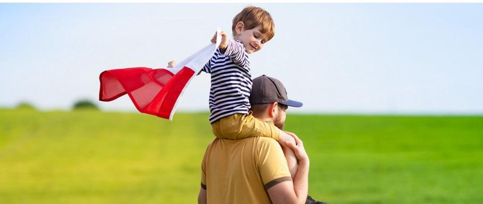 Mężczyzna trzyma na ramionach chłopca, który trzyma w dłoniach flagę biało-niebieską