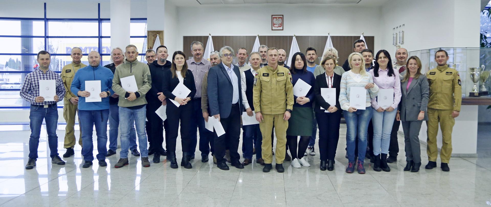 Grupowe zdjęcie uczestników szkolenia (23 osoby) w towarzystwie Zastępcy Komendanta Centralnej Szkoły PSP, dwóch funkcjonariuszy CS PSP oraz kobiety
