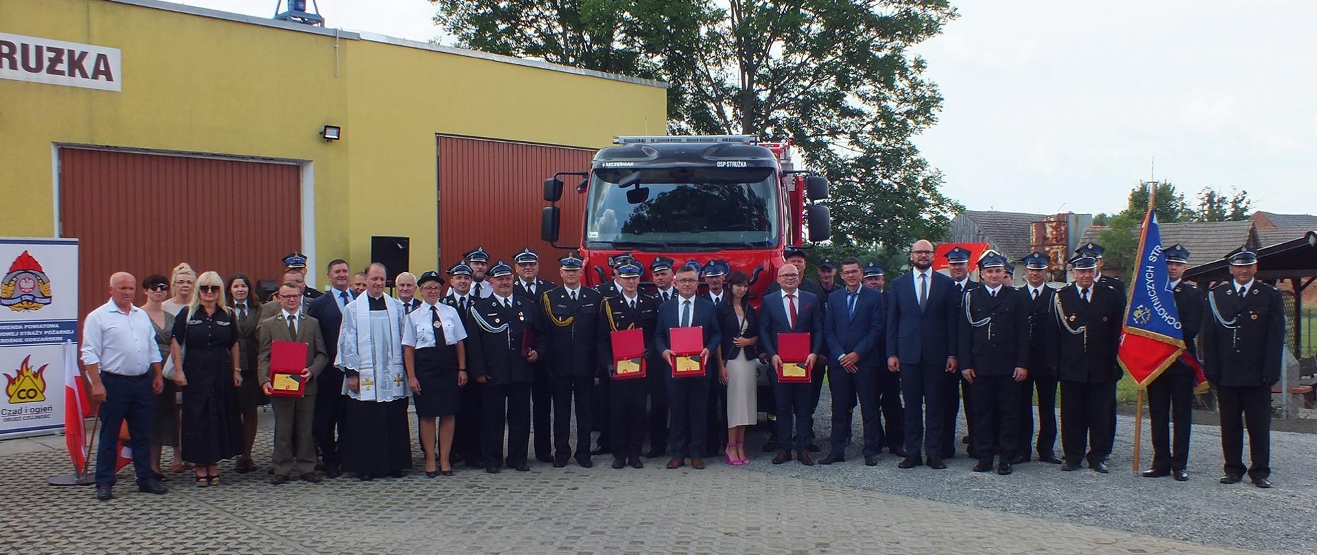 Zdjęcie przedstawia wszystkie zgromadzone osoby podczas przekazania nowego samochodu ratowniczo-gaśniczego. Zdjęcie wykonane zostało na tle remizy strażackiej oraz nowego samochodu.