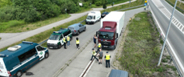 Inspektorzy WIOŚ w Warszawie kontrolują ciężarówkę. Kierowca otwiera drzwi naczepy, wewnątrz widać liczne pojemniki. Na poboczu stoją samochody współpracujacych służb.