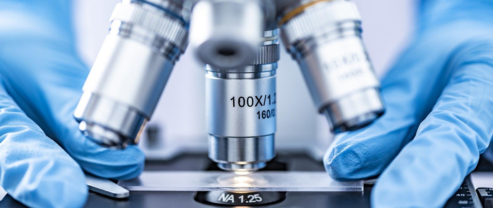 dłonie w niebieskich jednorazowych rękawiczkach kładą szkiełko pod mikroskop z materiałem do analizy