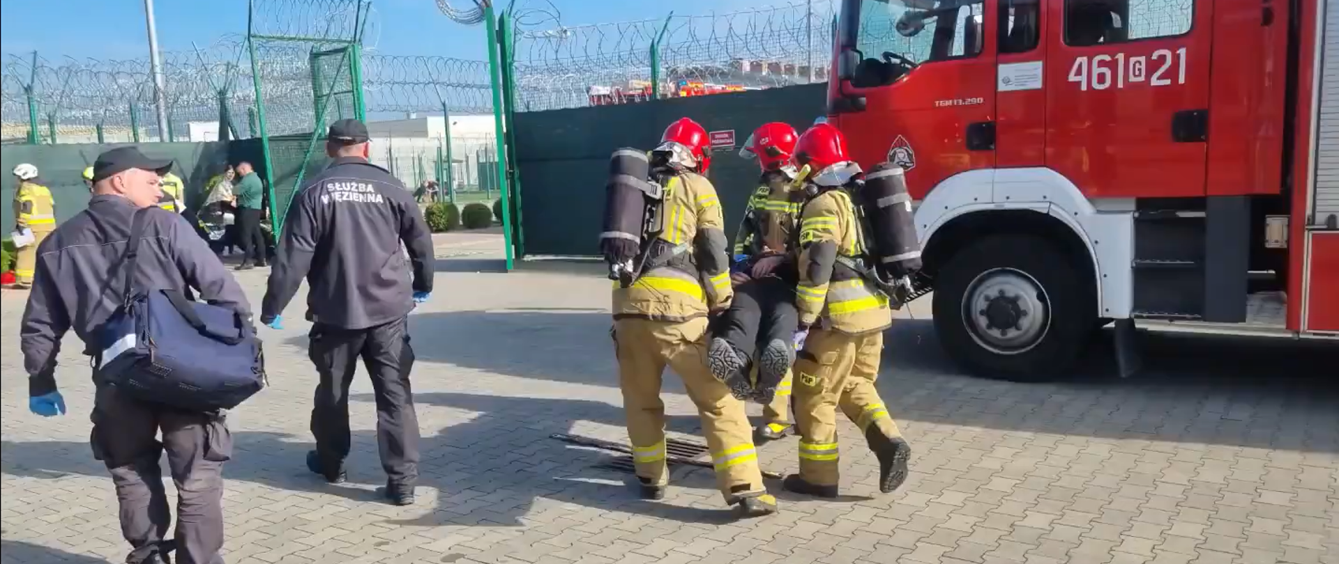 Strażacy przenoszą osobę poszkodowaną, obok funkcjonariusze Służby Więziennej 