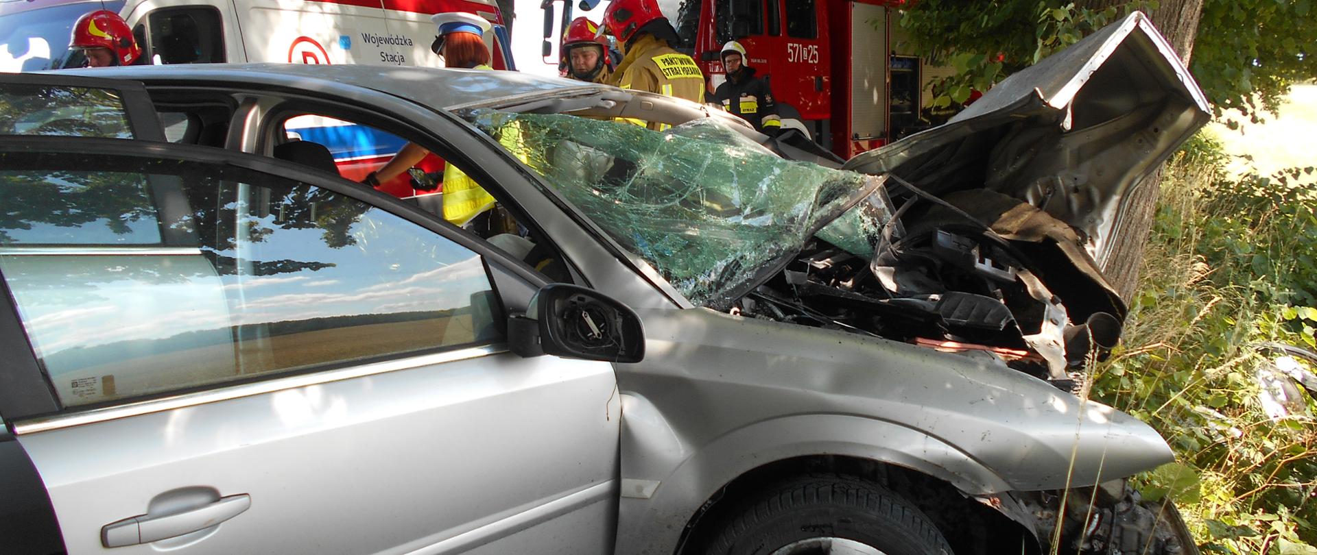 Zdjęcie przedstawia strażaków i samochód osobowy po uderzeniu w drzewo na tle wozu strażackiego i karetki pogotowia 