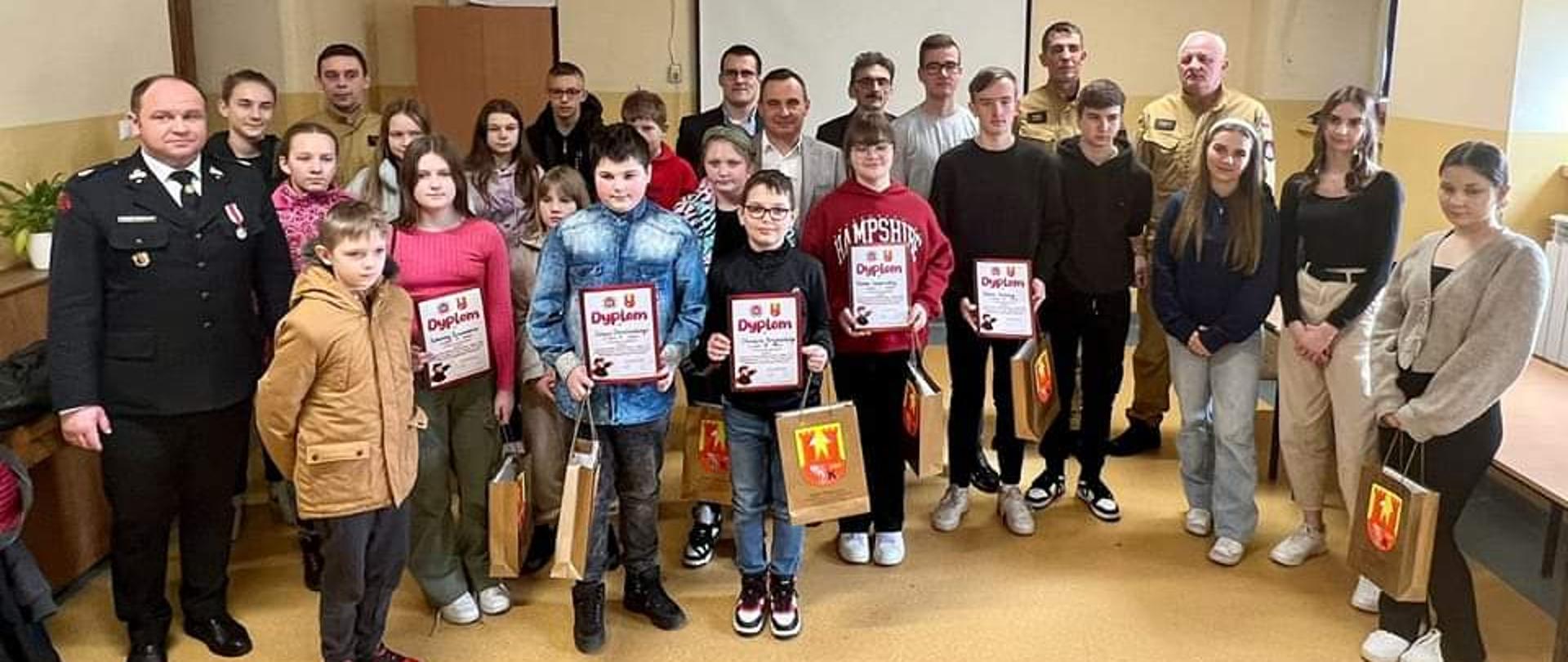 Sala w obiekcie w którym przeprowadzono etap gminny eliminacji, w pierwszych rzędach stoją uczestnicy turnieju oraz laureaci, w rękach trzymają dyplomy oraz nagrody, w tylnych rzędach przedstawiciele ZOSP RP, pracownicy gminy odpowiedzialni za ochronę przeciwpożarową, opiekunowie, władze gminy 