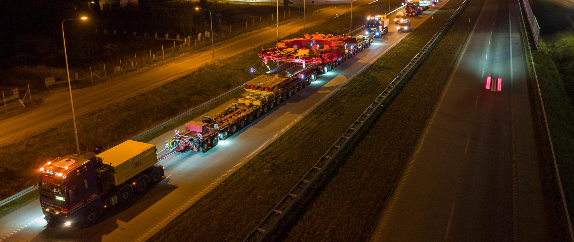 Na zdjęciu widoczny jest transport elementu maszyny do drążenia tunelu. Konwój składa się z kilku dużych ciężarówek, które przewożą masywne części maszyny. Transport odbywa się nocą, co widać po oświetleniu ulicznym i ciemnym niebie. Droga jest dobrze oświetlona, a pojazdy są wyposażone w światła ostrzegawcze. W tle można dostrzec stację benzynową oraz inne pojazdy uczestniczące w konwoju.