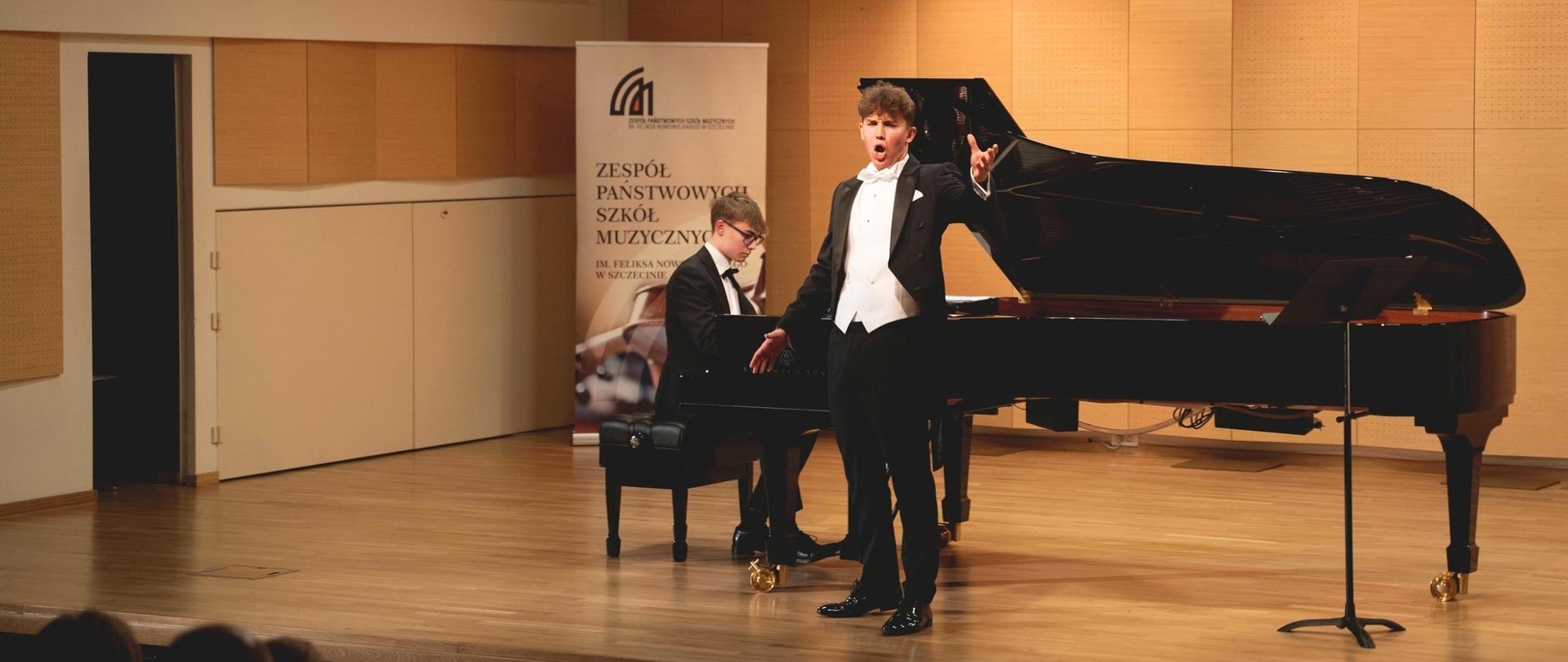 zdjęcie przedstawia ucznia śpiewającego z towarzyszeniem pianisty grającego na otwartym fortepianie na scenie sali koncertowej, w tle baner szkoły, inne ujęcie z szerszej perspektywy
