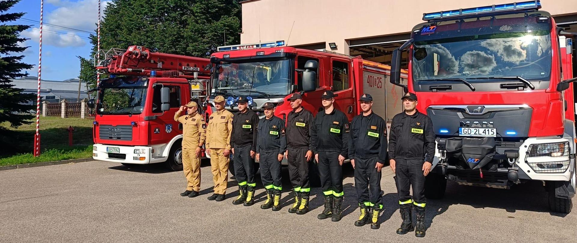 Strażacy w szeregu stoją na tle pojazdów pożarniczych 