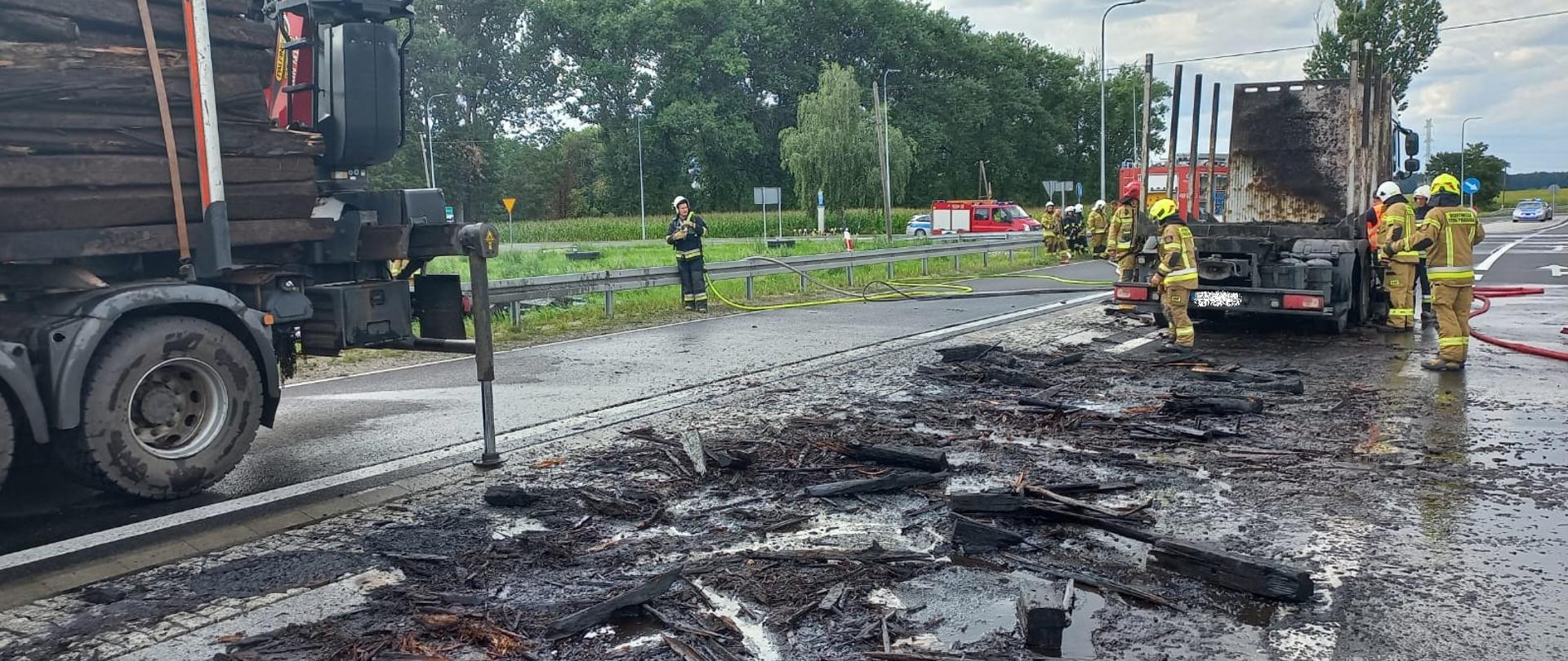 Po prawej stronie stoi samochód ciężarowy który przewoził podkłady. Za nim resztki podkładów, które spadły na drogę podczas rozładunku. Po lewej stronie drugi pojazd przewożący podkłady. Obok samochodów stoją strażacy.
