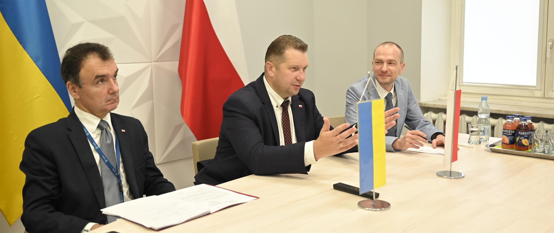 Minister Czarnek i dwóch mężczyzn siedzą przy stoliku, na którym stoją małe flagi Polski i Ukrainy.