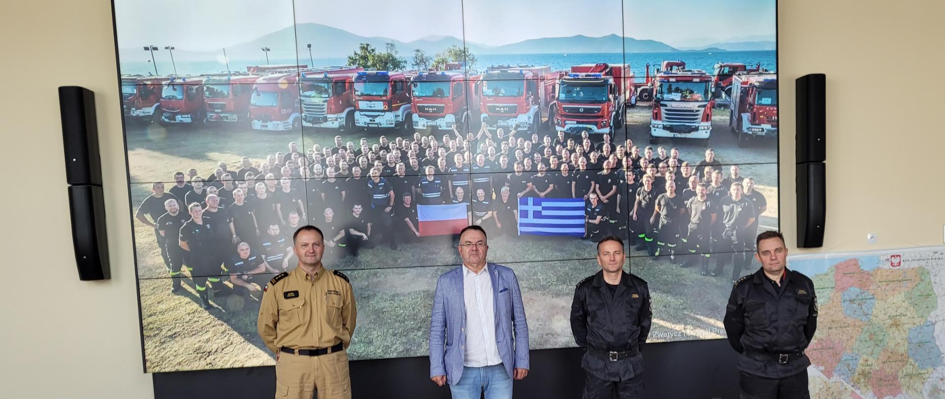 Pozują do zdjęcia (od lewej) Dyrektor KCKRiOL , Prezes Izby i dwóch zastępców dyrektora KCKRiOL. W tle wyświetlone zdjęcie polskich strażaków w Grecji