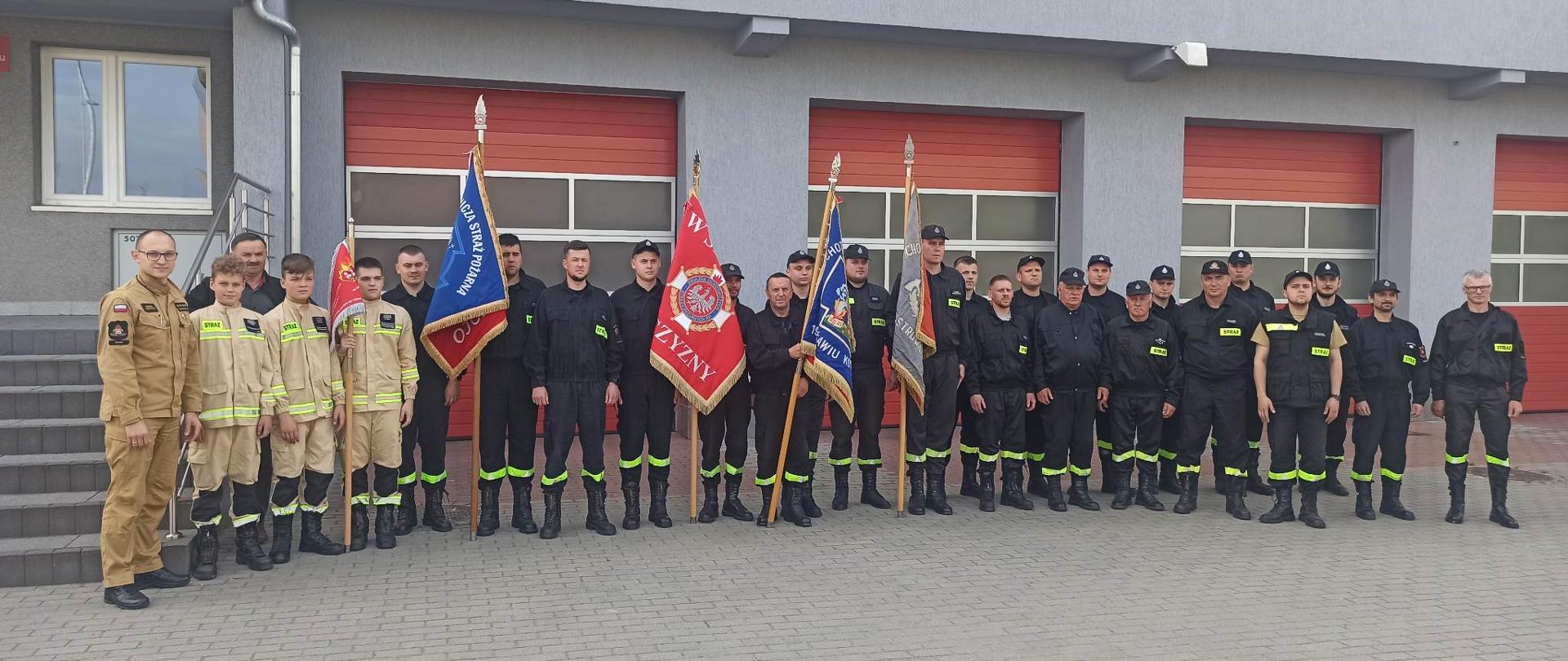 Zdjęcie przedstawia strażaków oraz druhów OSP stojących na placu przed budynkiem komendy podczas szkolenia pocztów sztandarowych. 
