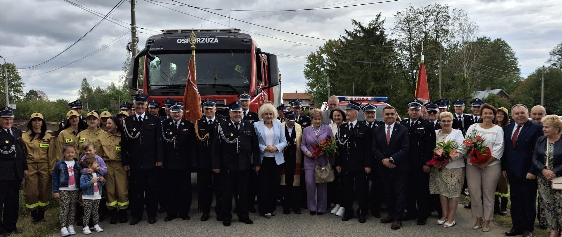 Zdjęcie grupowe uczestników kilkudziesięciu uroczystości pozujących na tle samochodów strażackich. Wśród nich kilku strażaków strażaków w mundurach galowych. W głębi widoczne są zielone korony drzew i pochmurne niebo