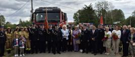 Zdjęcie grupowe uczestników kilkudziesięciu uroczystości pozujących na tle samochodów strażackich. Wśród nich kilku strażaków strażaków w mundurach galowych. W głębi widoczne są zielone korony drzew i pochmurne niebo