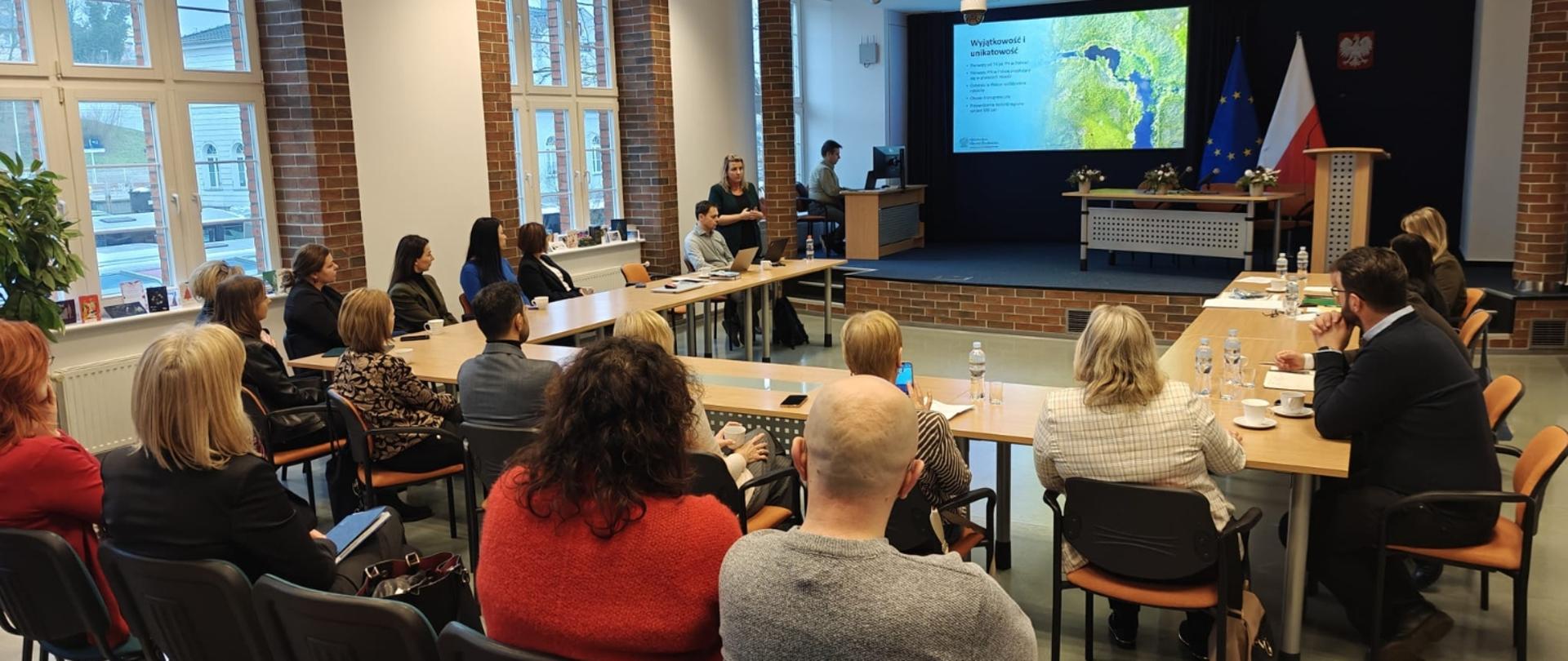 Osoby zgromadzone przy stołach w sali podczas spotkania konsultacyjnego, uczestniczące w dyskusji i interakcji społecznej.