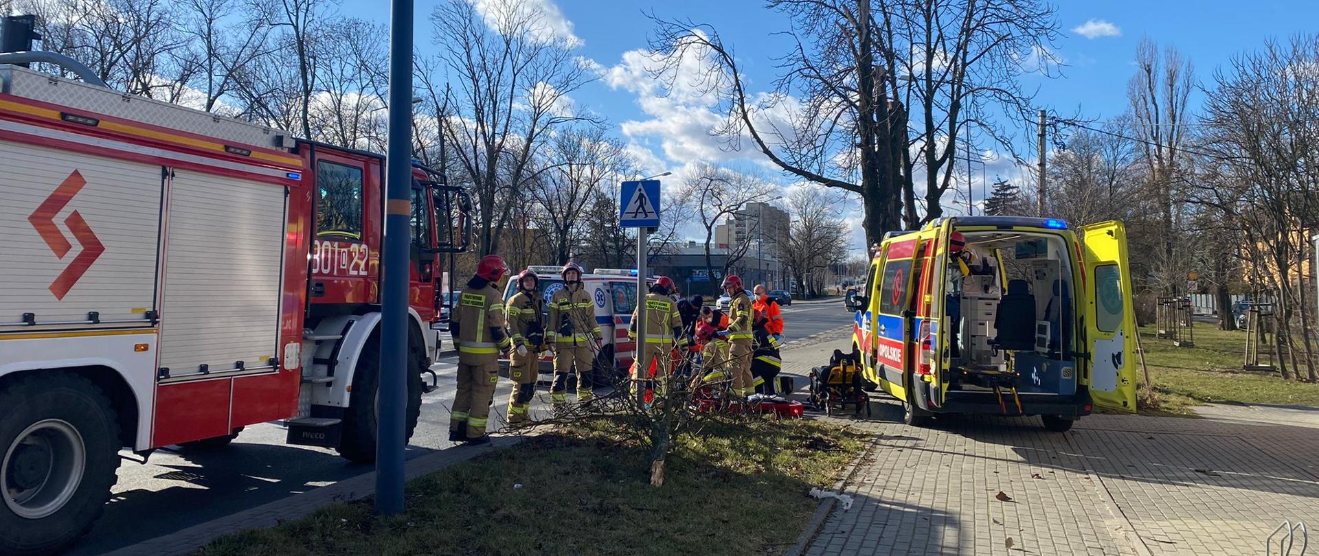 Strażacy oraz ratownicy medyczni udzielają pomocy osobie poszkodowanej, która została uderzona spadającym konarem drzewa. Po lewej widoczny samochód pożarniczy koloru czerwonego a po prawej ambulans zespołu ratownictwa medycznego koloru żółtego. Strażacy w umundurowaniu specjalnym koloru piaskowego z czerwonymi hełmami na głowach.