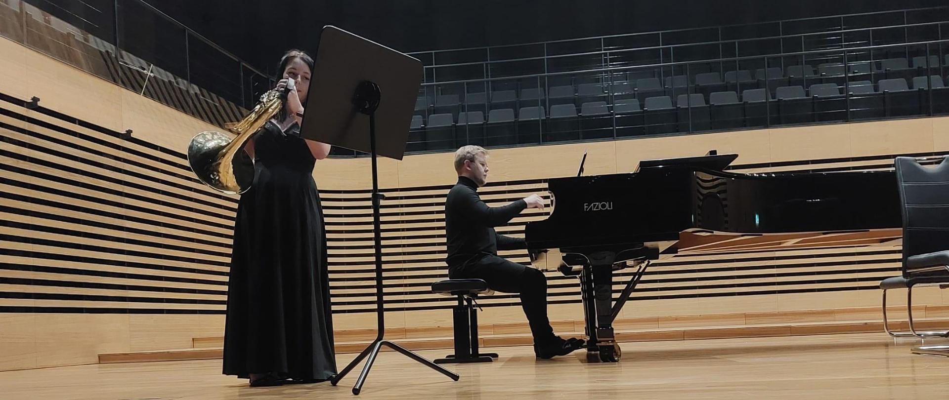 Nastolatka stoi przy pulpicie na etsradzie sali koncertowej i gra na waltorni, za nią na fortepianie Fazioli gra mężczyzna.