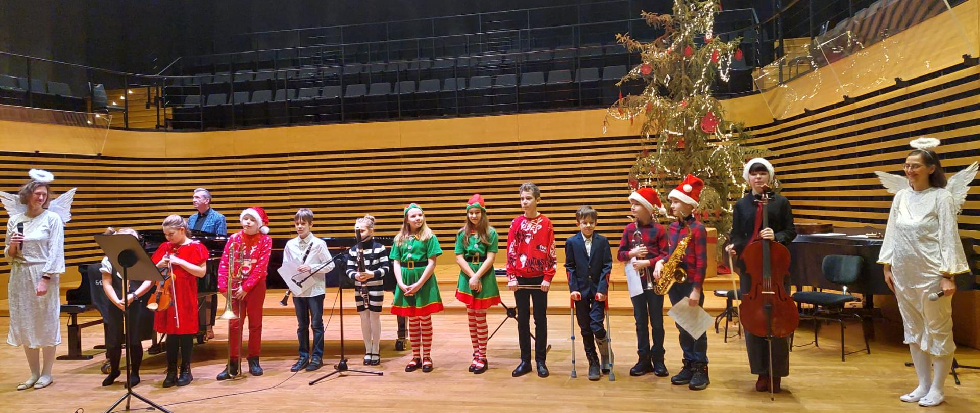 Wszyscy wykonawcy stoją ze swoimi instrumentami na scenie, po bokach dwie kobiety w strojach aniołków, z tyłu stoi mężczyzna.