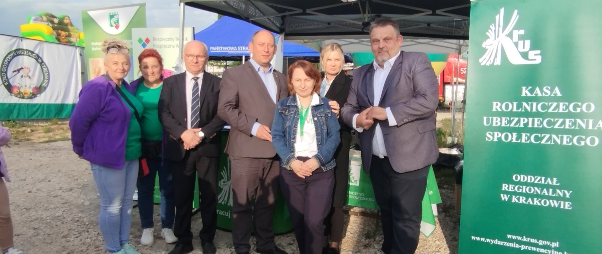 Grupa osób, cztery kobiety i trzech mężczyzn pozuje do wspólnego zdjęcia przy stoisku informacyjnym KRUS podczas Pikniku Rodzinnego. W tle widoczne namioty oraz baner Kasy Rolniczego Ubezpieczenia Społecznego Oddział Regionalny w Krakowie.