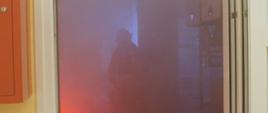 Na zdjęciu widoczne jest zadymione pomieszczenie, w którym strażacy szukają osób poszkodowanych. Wewnątrz widać światła imitujące płomienie.