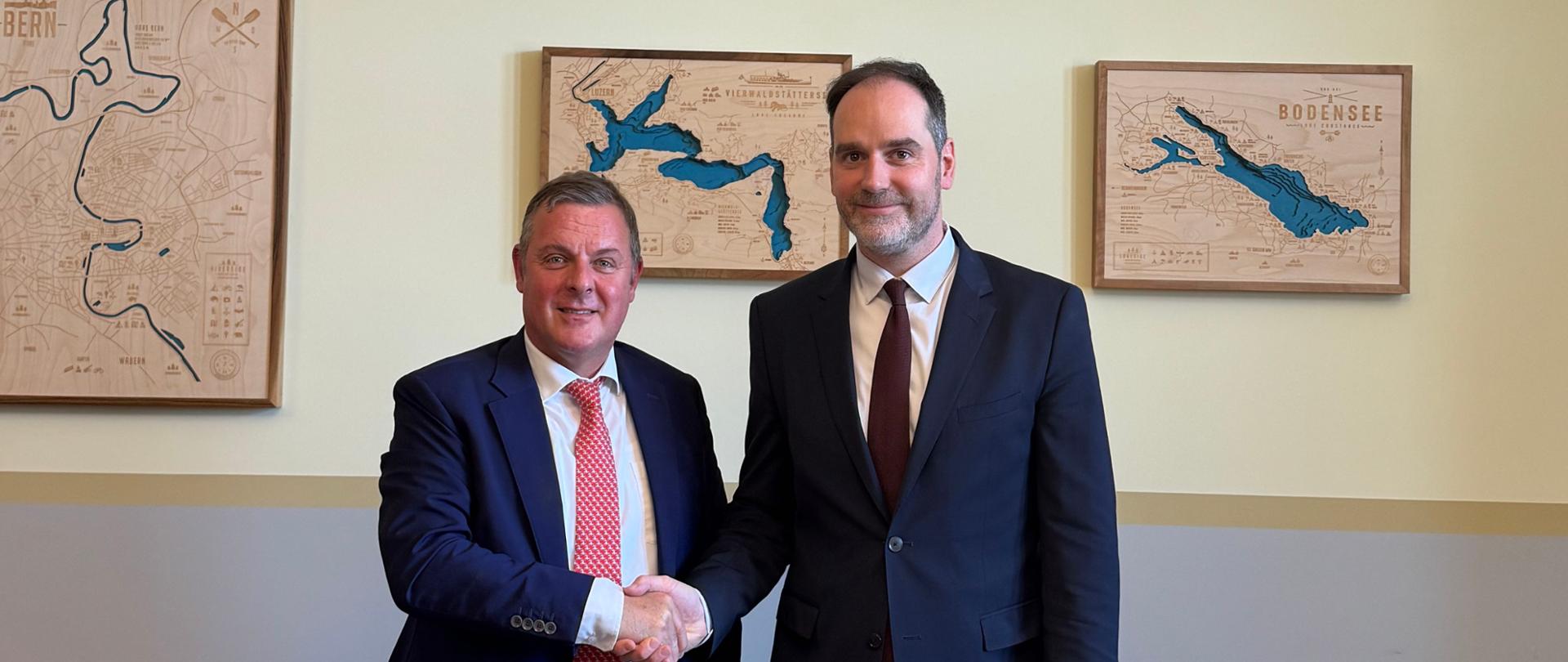 Deputy Minister Ignacy Niemczycki shakes hands with Patric Franzen during an official meeting. On the table in front of them are small flags—those of Switzerland and Poland. Various maps are visible on the wall in the background.