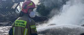 Widok z boku. Strażak KP PSP w Wadowicach ubrany w odzież specjalną, hełm i maskę przeciwpyłową podaje prąd wody na stertę słomy celem jej dogaszenia. 
