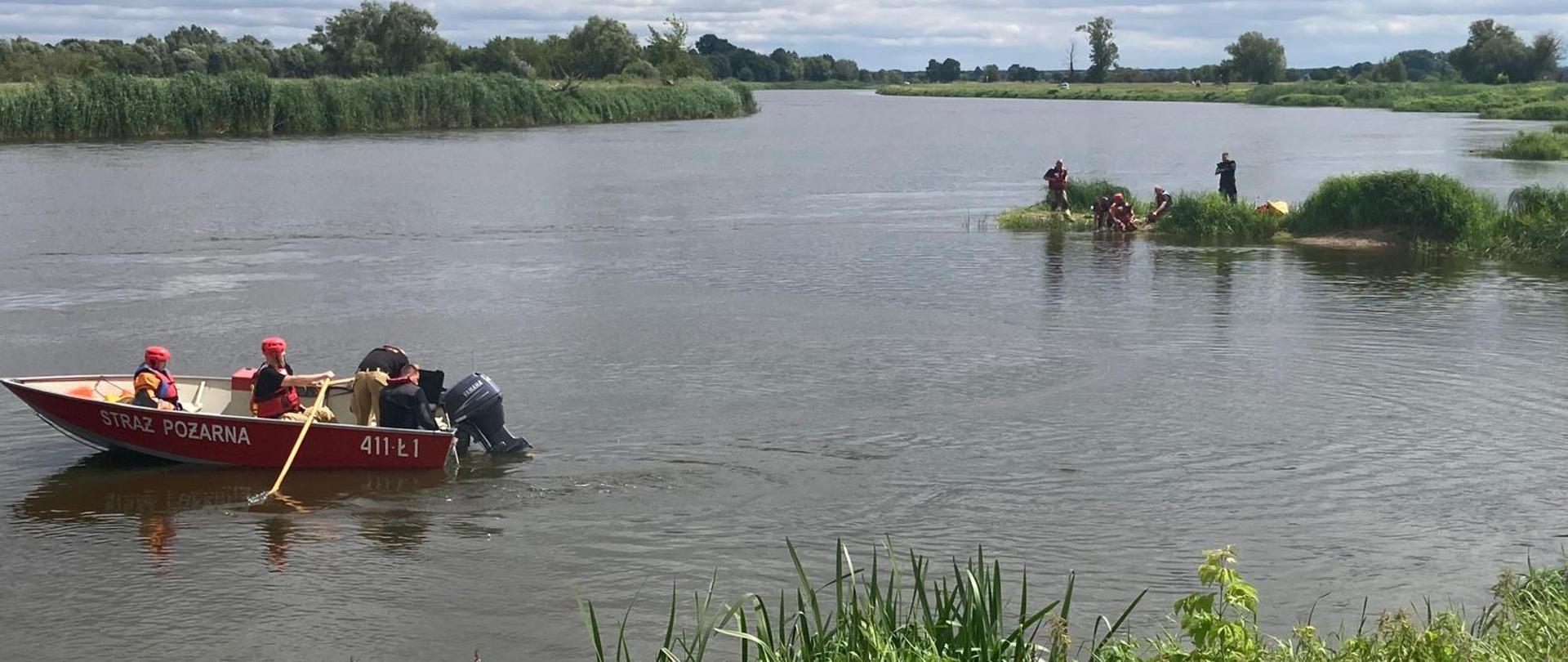Strażacy płynący łodzią, w oddali strażacy stojący na brzegu rzeki