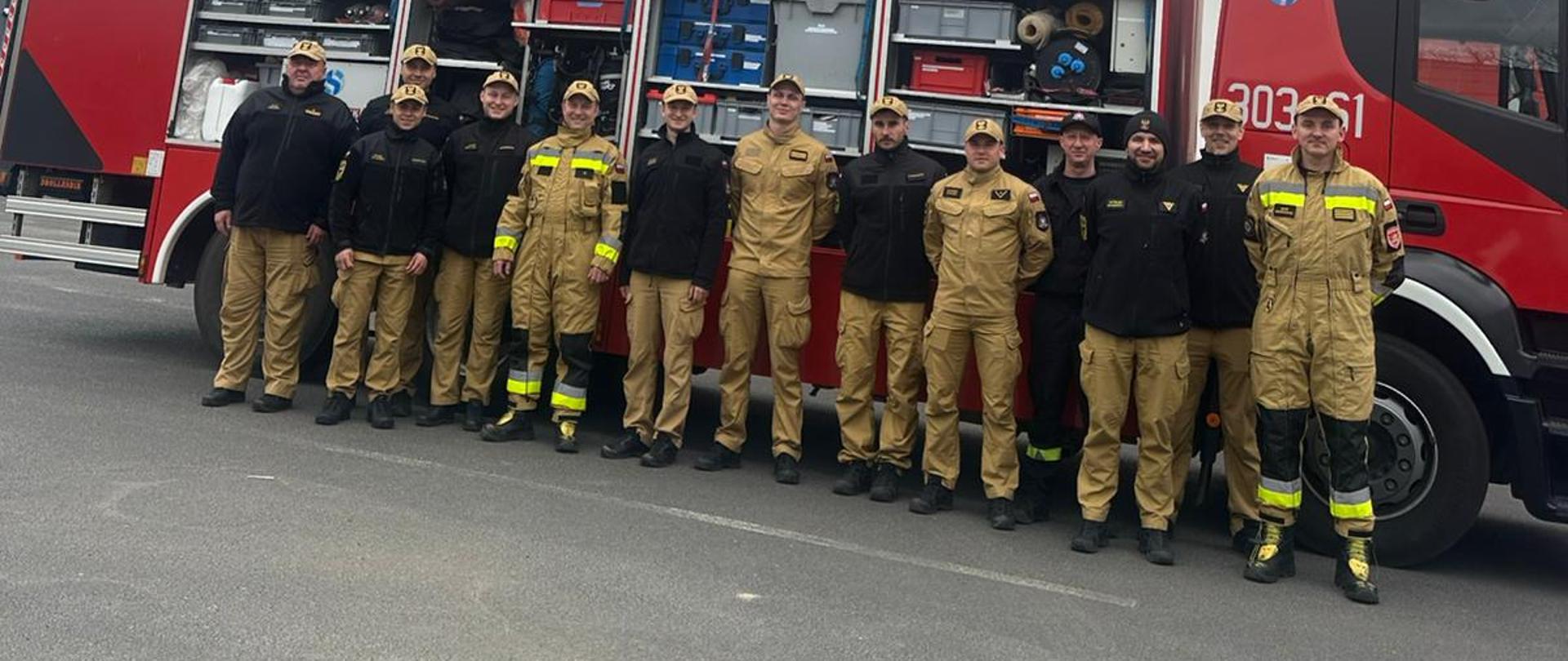 Zdjęcie przedstawia grupę kilkunastu strażaków pozujących na tle nowoczesnego, czerwonego wozu strażackiego. Strażacy stoją w jednym rzędzie; mają na sobie piaskowe i czarne mundury służbowe oraz czapki z daszkiem. Boczne skrytki wozu strażackiego są otwarte, ukazując specjalistyczny sprzęt ratowniczy ułożony na półkach. W tle widoczny jest biało-czerwony budynek remizy pod zachmurzonym niebem.